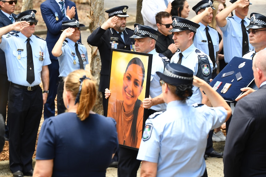 a procession of police officers along a street holding a large framed photo of a young officer who was shot dead