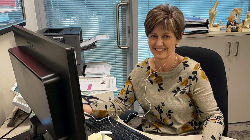 Woman sits behind computer, holding mouse and wearing headphones