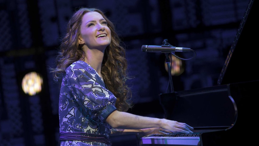 Esther Hannaford, performing as Carole King, sits at a grand piano on stage and smiles towards the audience.