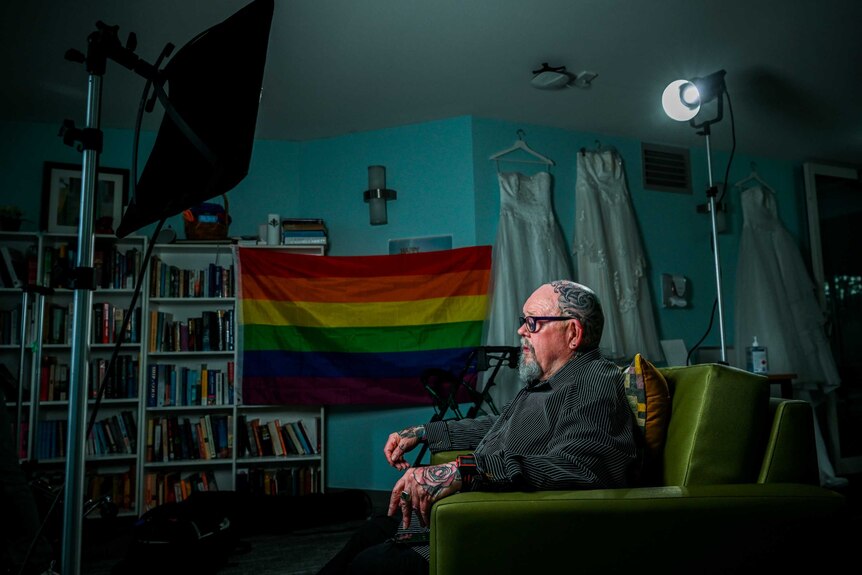 Geoffrey Ostling sit in his room in front of a rainbow flag, books line one wall, while wedding dresses hang on another.