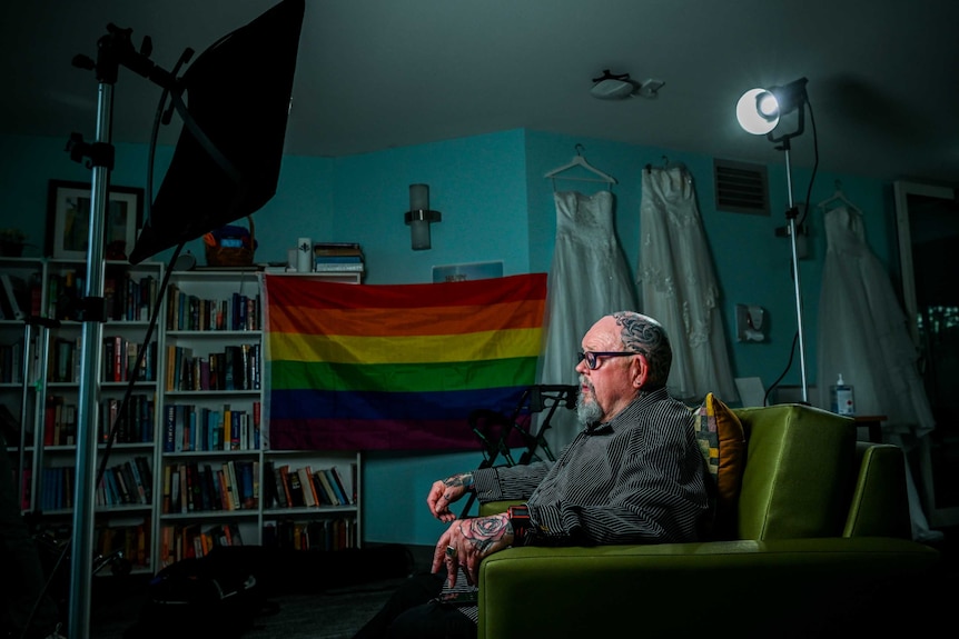 Geoffrey Ostling sit in his room in front of a rainbow flag, books line one wall, while wedding dresses hang on another.