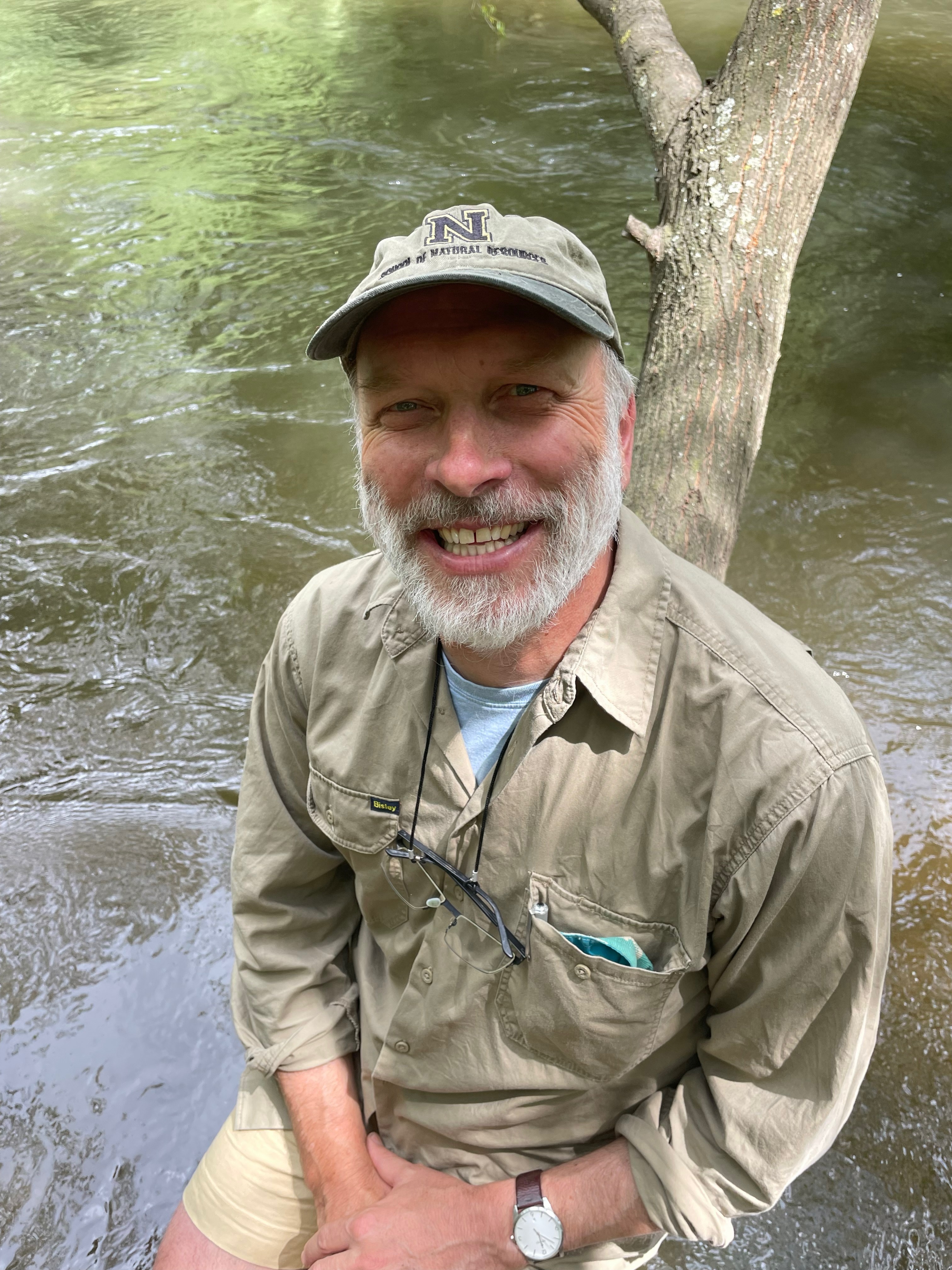 A smiling older bearded man sitting on a tree in front of a body of water.
