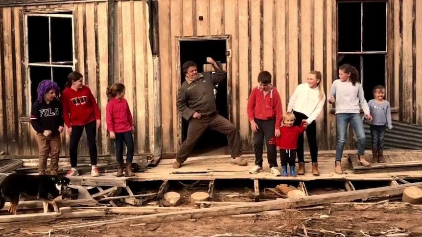 A middle aged man and several children dance outside an old timer shed in a dusty landscape.