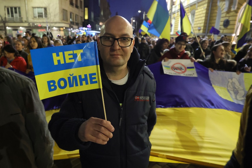 A bald man in glasses stands in front of a sea of people holding a small plastic flag that reads 'no war' in Ukrainian.