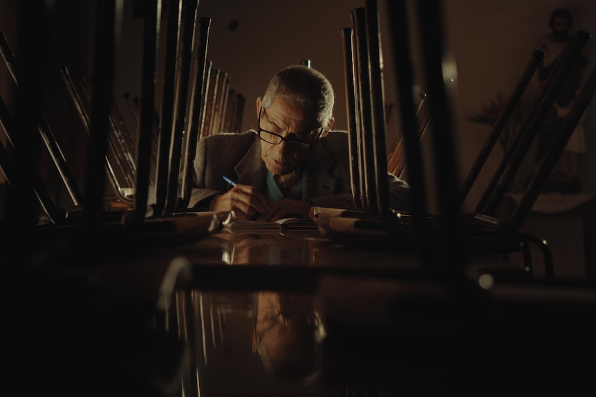 An elderly man, the spy Sergio Chamy, writing notes surrounded by upturned chairs in documentary The Mole Agent