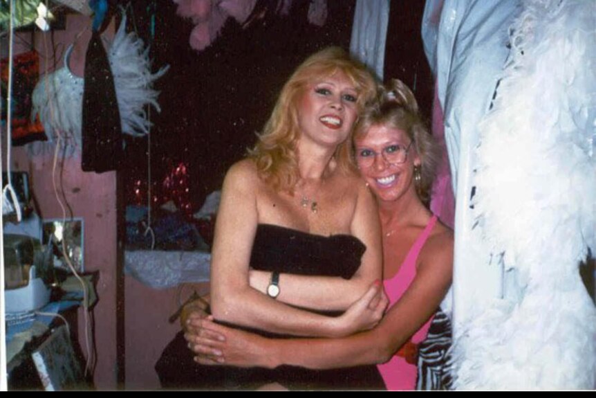 A transgender woman with blonde hair sitting on the lap of another transgender woman with blonde hair.