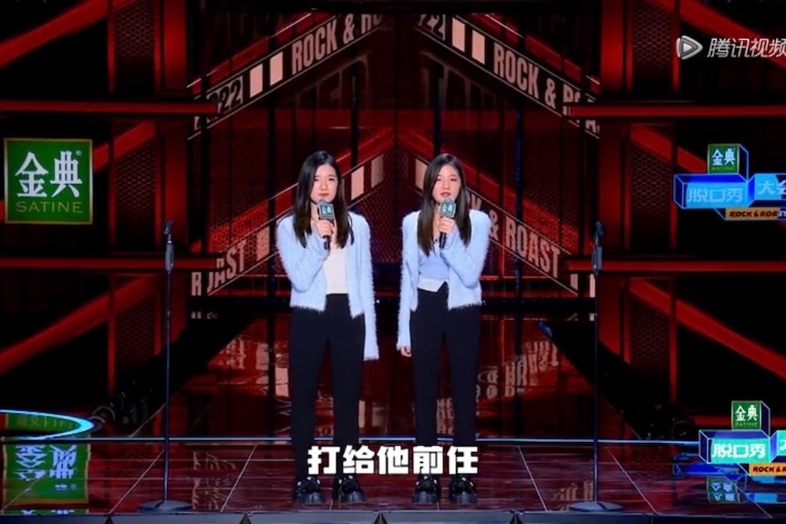 Female Chinese twins dressed in identical clothes speak on a stage.
