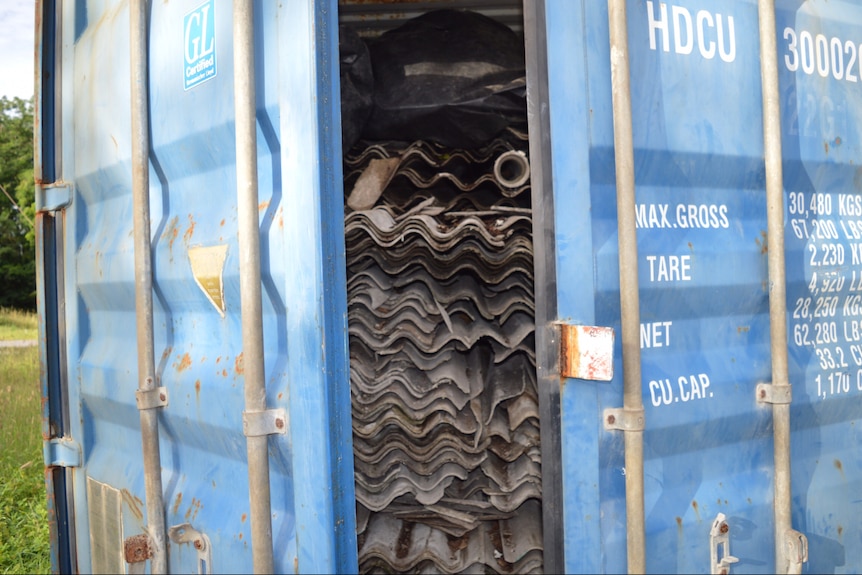 A blue shipping container's door is open, and sheets of asbestos are packed to the top.