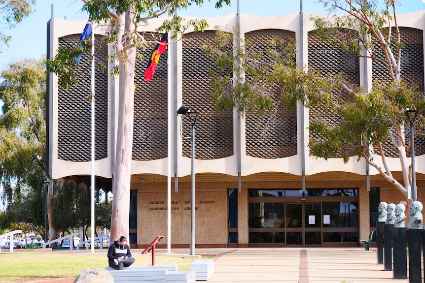 A young girl wearing a black hoodie sits on a bench in front of Broken Hill City Council