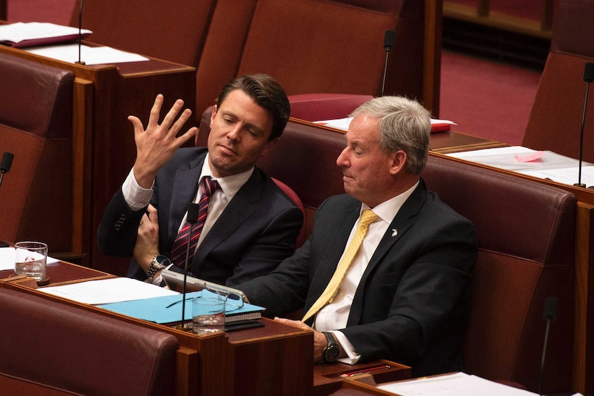 Two men in suits sit alone in the red Senate chamber