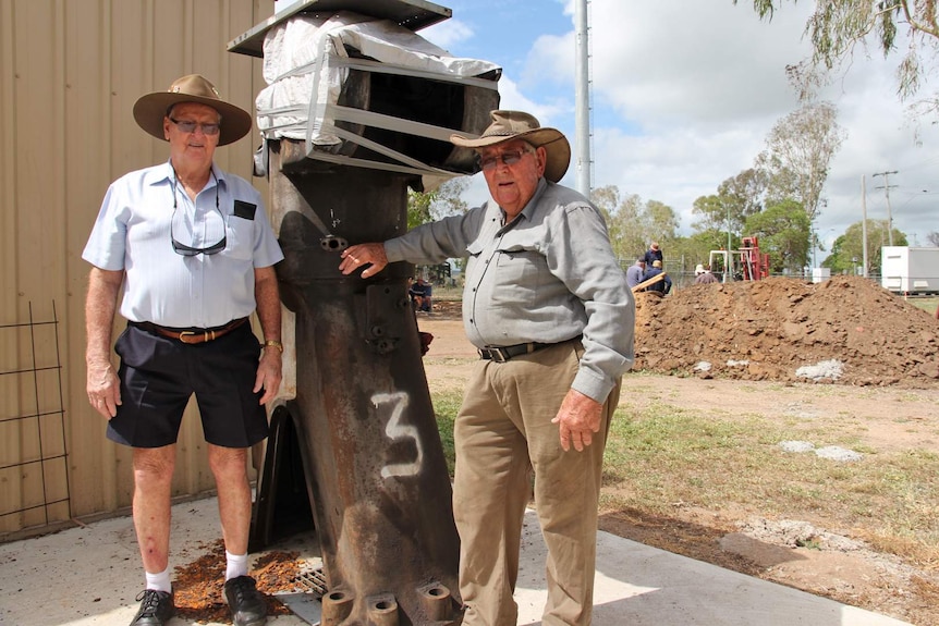 Two men stand with a cylinder from a massive vintage engine removed from a sugar mill