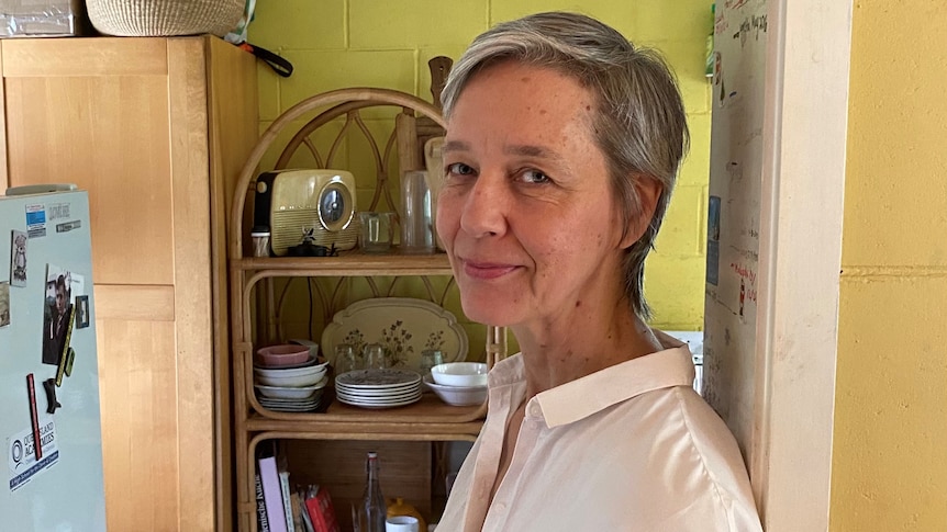 A woman with short grey hair stands in front of a kitchen shelf