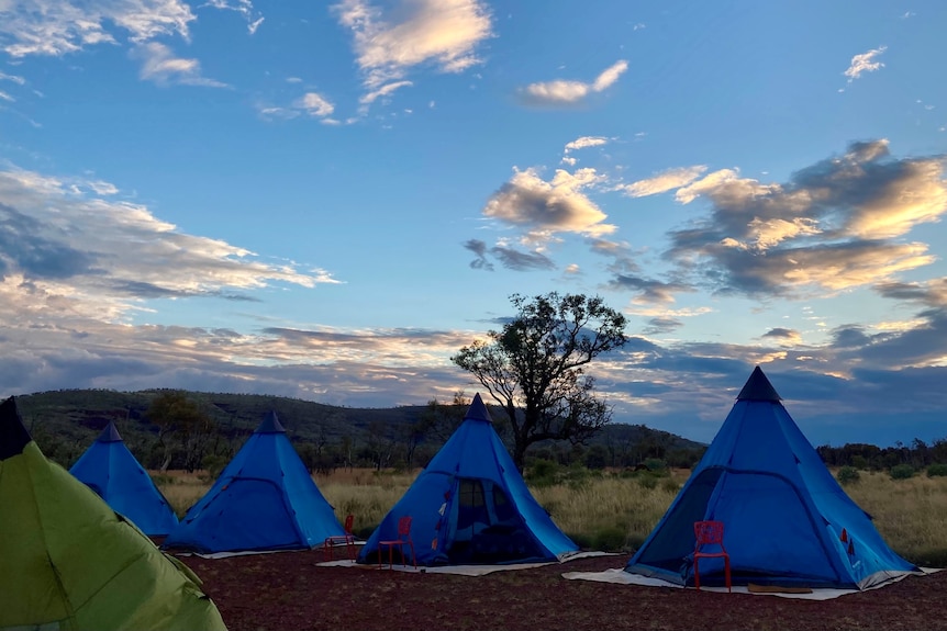 A row of tents stands on a scrubby plain as the suns sets  behind nearby hills.