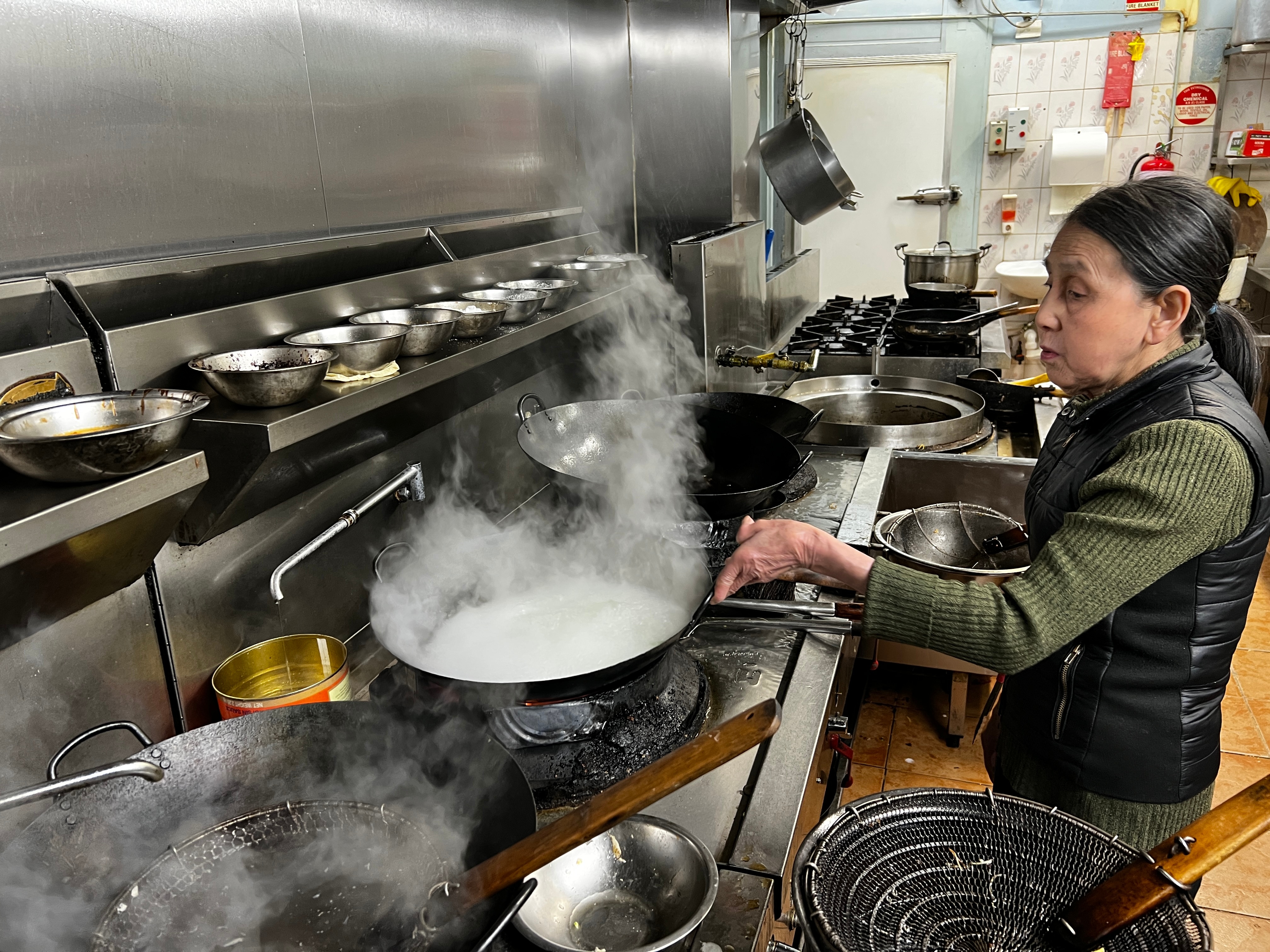 An older Vietnamese woman cooks with gas on a wok in a kitchen 