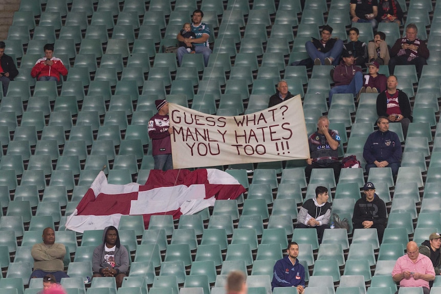 Manly fans hold a sign reading 'GUESS WHAT? MANLY HATES YOU TOO!!!' in the stands before a game.