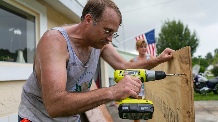 A man leans in to drill a hole through plywood to board up a window