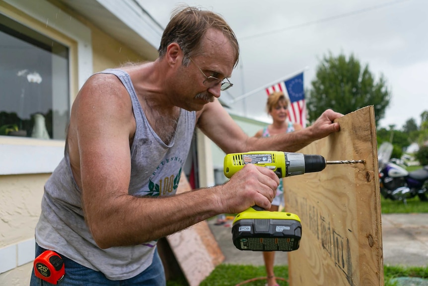 A man leans in to drill a hole through plywood to board up a window