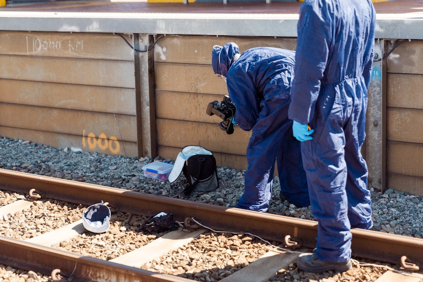 Two WA Police forensic officers in protective clothing take photographs of a bag and other items on train tracks.