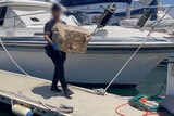 Police officer walking along a marina carrying a large box wrapped in brown paper 