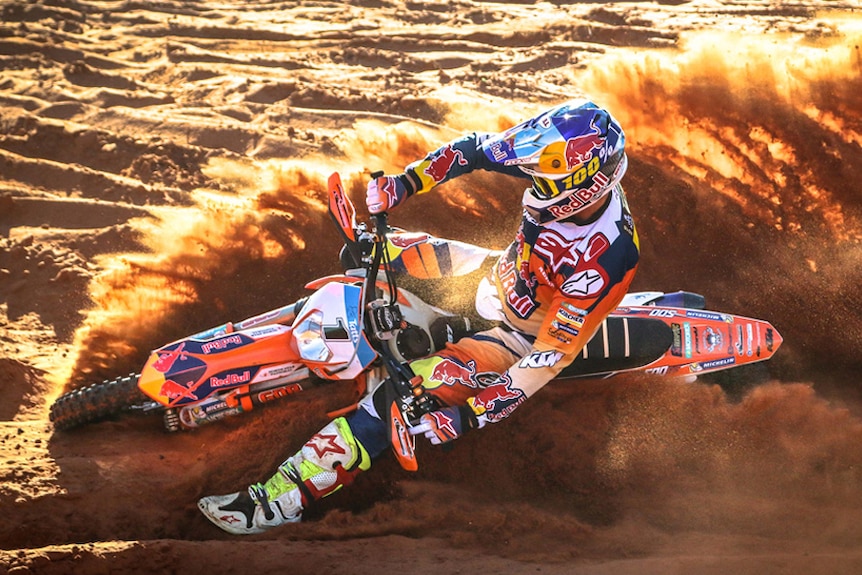A man in colourful motorbike racing gear skids in the red dirt, sending up a cloud of dust.