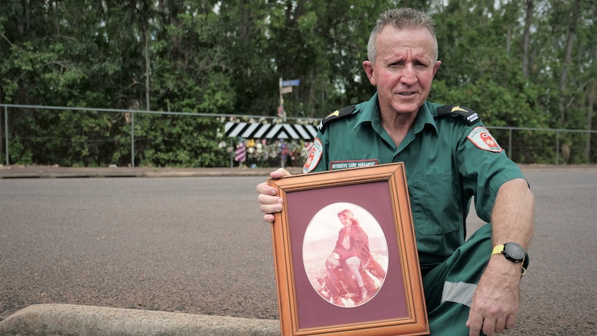 Man wearing paramedic uniform sitting on kerb opposite roadside memorial holding photo of woman.