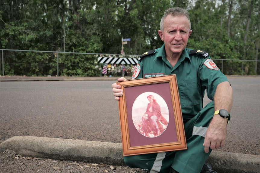 Man wearing paramedic uniform sitting on kerb opposite roadside memorial holding photo of woman.