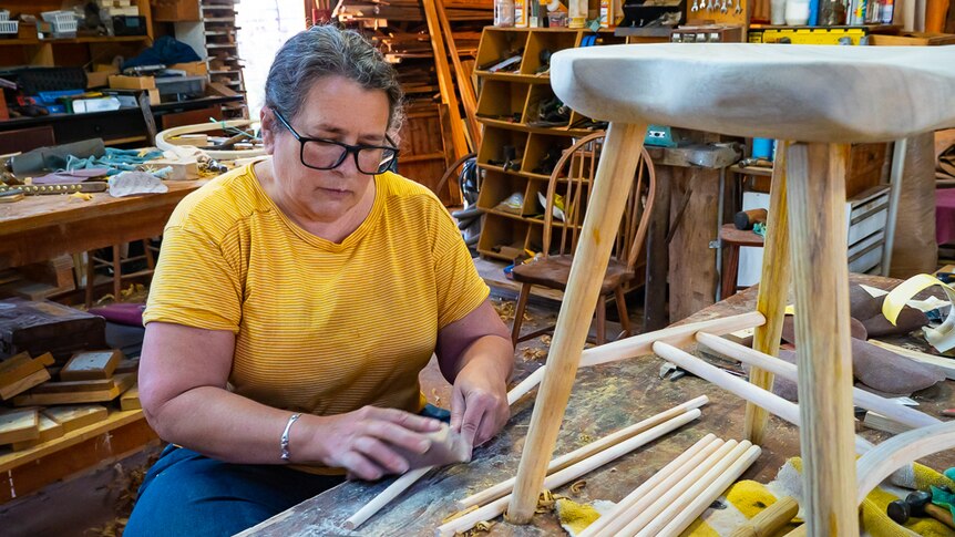 A woman sanding wooden rods with a chair in the foreground.