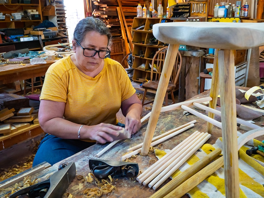 A woman sanding wooden rods with a chair in the foreground.