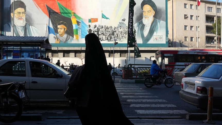 A woman, wearing the chador, a head-to-toe garment, walks on a sidewalk in front of a mural of Iran leaders.