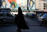 A woman, wearing the chador, a head-to-toe garment, walks on a sidewalk in front of a mural of Iran leaders.