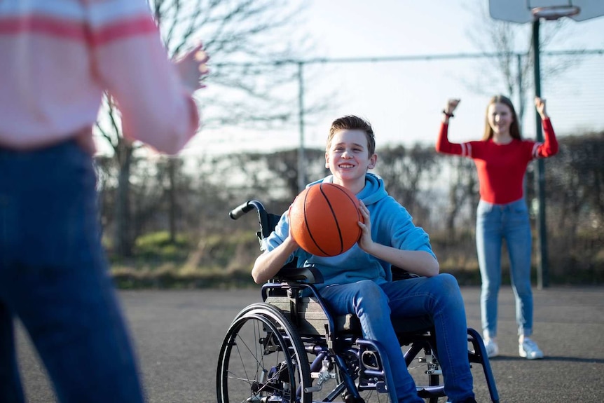 A primary school boy in a wheelchair is about to throw a basketball to another student. They are on an outdoor basketball court.