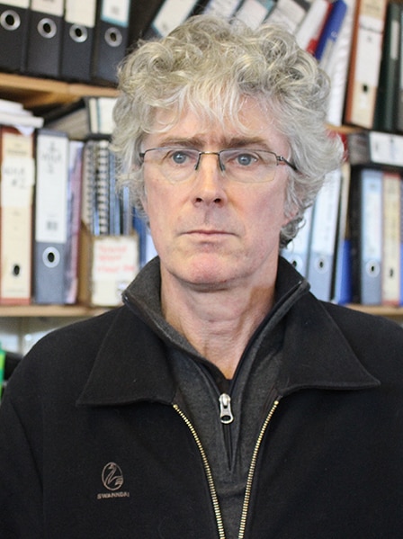 Peter McGlone, looking serious, stands in front of a book shelf.