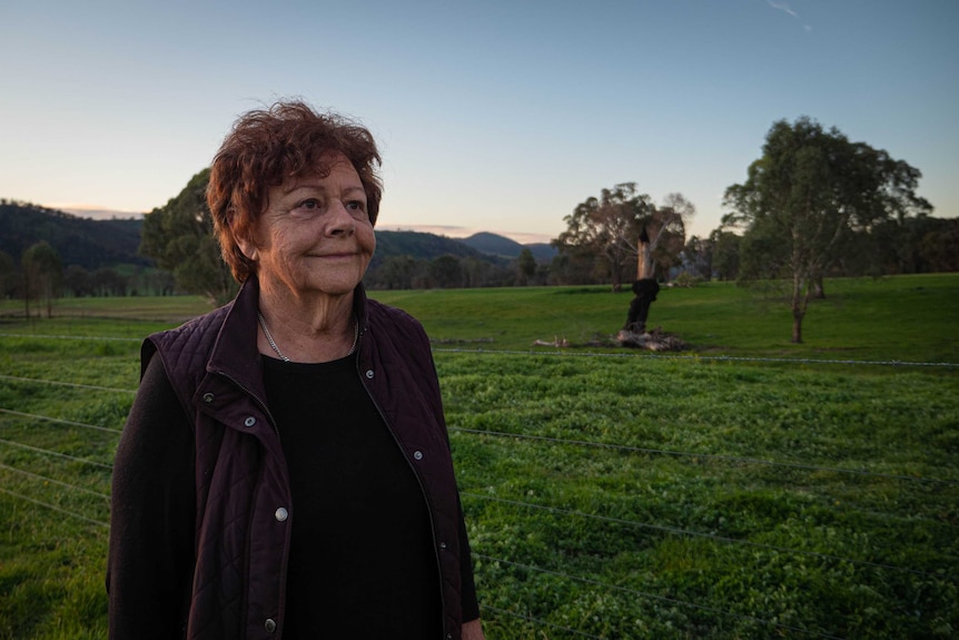 A woman with amber hair, wears a black top and dark purple vest, she's standing in a green paddock with a burnt tree behind.