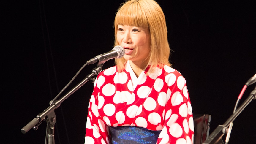 A woman in a kimono speaks into a microphone, black background.