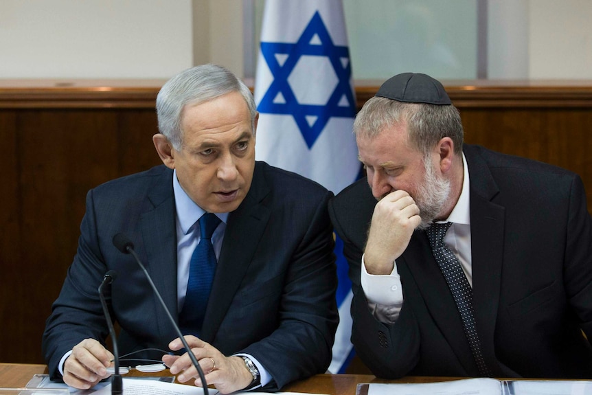 Benjamin Netanyahu speaks with Avichai Mandelblit, who is wearing a black kippah, with an Israeli flag in the background