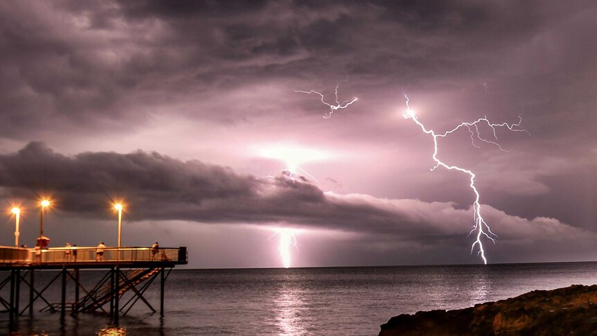 Lightning strikes stand out against a purple, cloudy sky in the distance, with people watching from a jetty on the water.