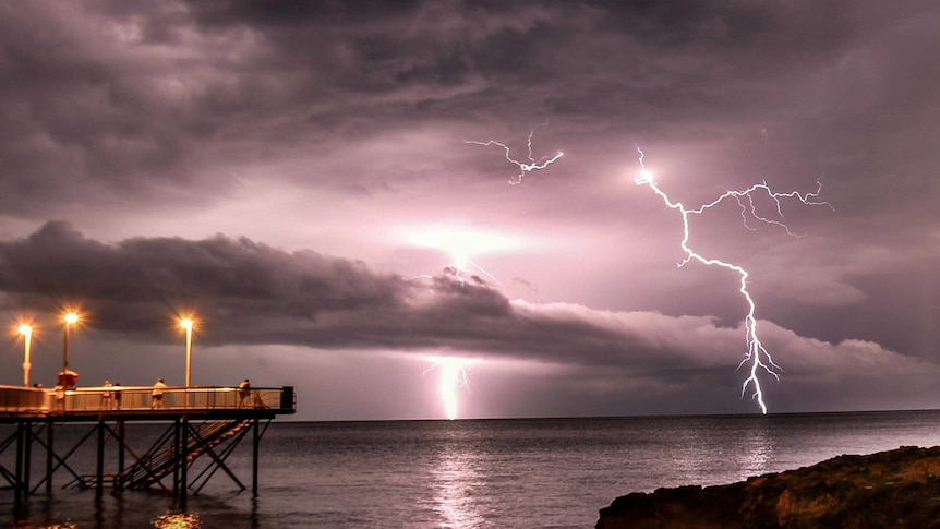 Lightning strikes stand out against a purple, cloudy sky in the distance, with people watching from a jetty on the water.