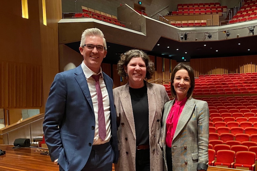 A man and two women standing next to each other on stage of empty auditorium.