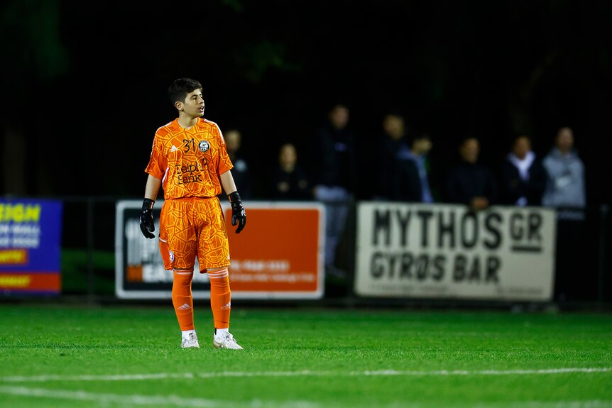 A boy stands in an orange goalkeeper's football kit during an Australia Cup match for Oakleigh Cannons.