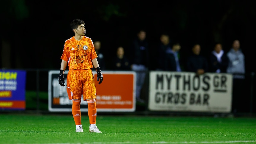 A boy stands in an orange goalkeeper's football kit during an Australia Cup match for Oakleigh Cannons.