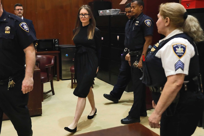 Woman in glasses wearing a black dress walks into a court room surrounded by guards with her hands handcuffed behind her back.
