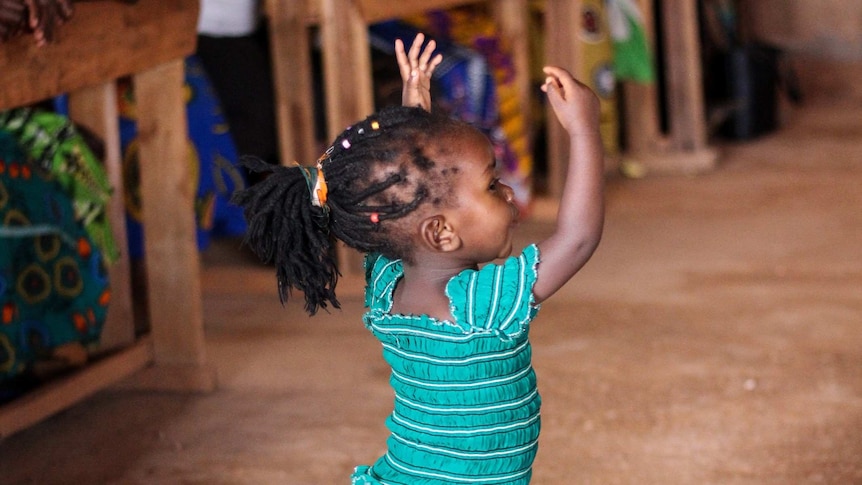 A photograph of a small child in a blue dress waving her arms above her head while dancing.