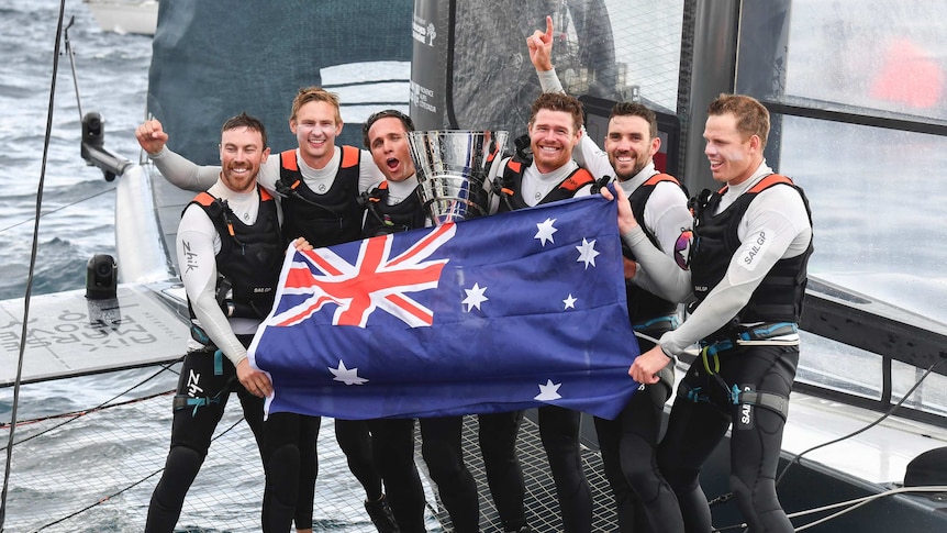 Team Australia celebrates with a large silver trophy and an Australia flag, standing on a boat
