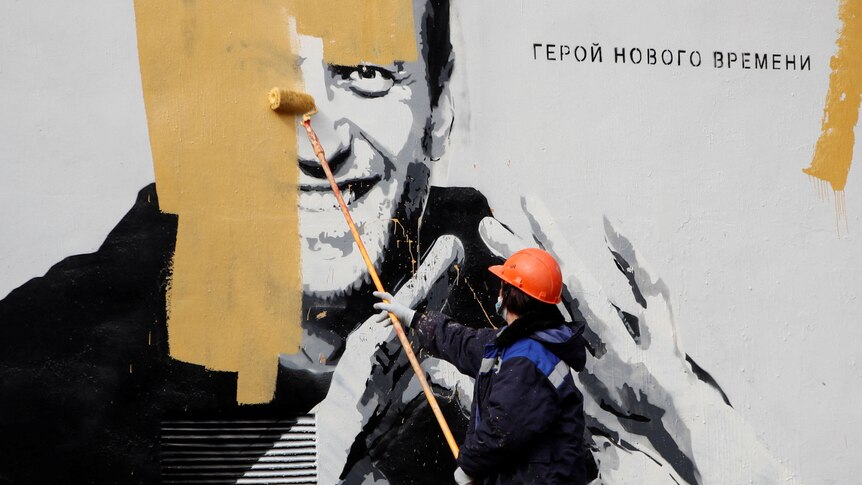 A person in winter clothing an an orange hard hat paints yellow paint over a large portrait of a man painted on a wall.