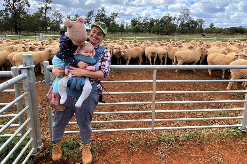 A smiling woman holds her two children in front of a pen teeming with sheep.