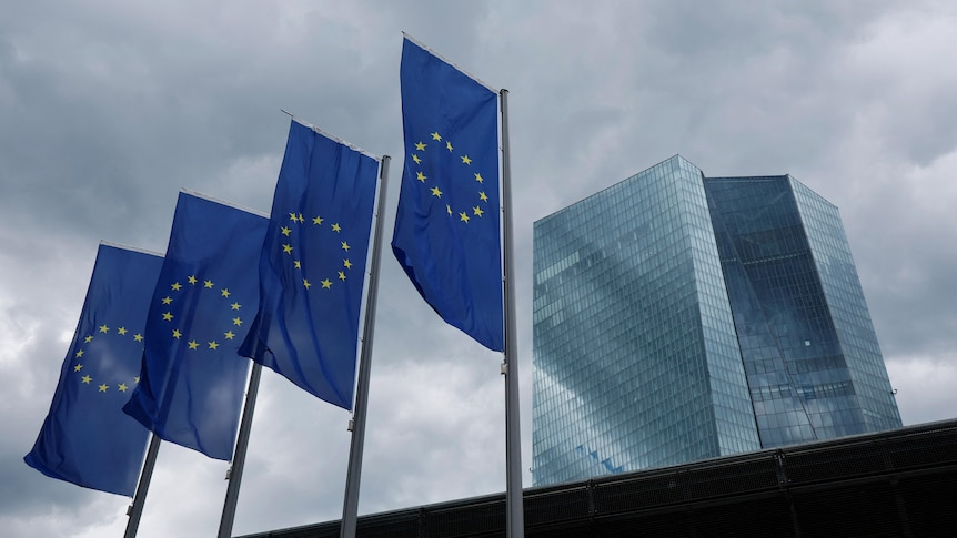 Four European Union flags fly outside a large tall corporate building on a dark and stormy day.