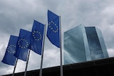 Four European Union flags fly outside a large tall corporate building on a dark and stormy day.