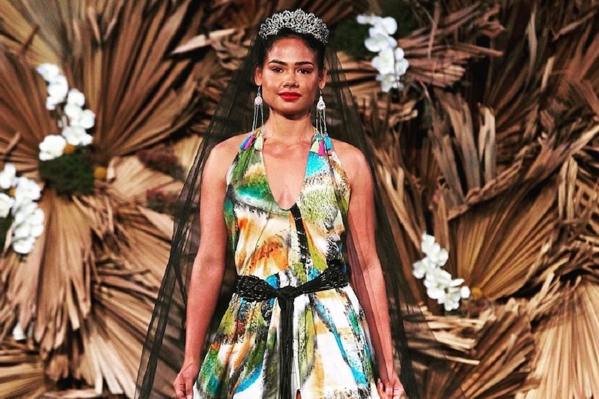 A polynesian woman in a long dress with green, black blue flowers walking down a runway with dry leaves in the background