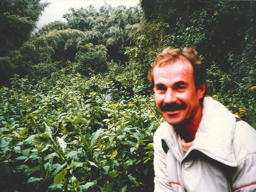 A man with a mistache in the forground smiles, in the background rainforest and gorillas