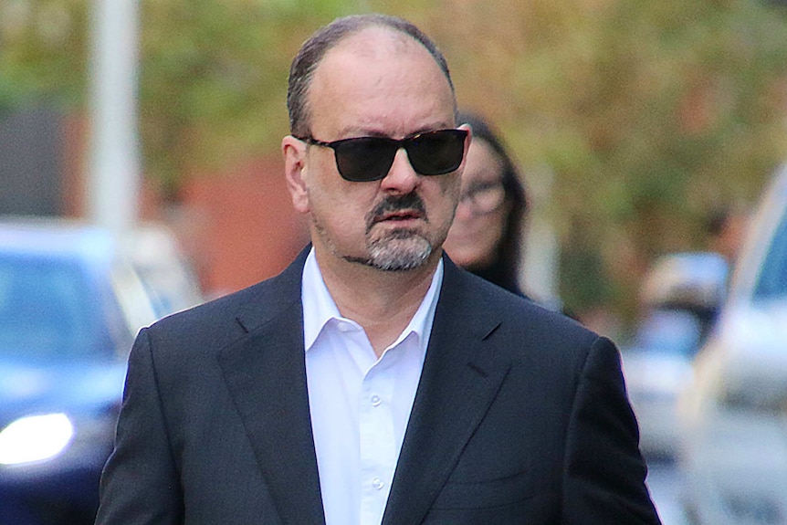 A mid-shot of lawyer Paul Yovich walking outside court in a black suit, white shirt and sunglasses.